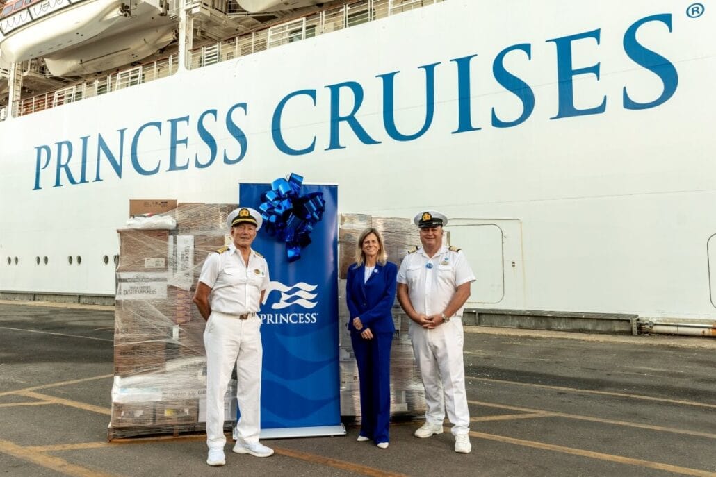 Three people stand in front of a large cruise ship with "Princess Cruises" written on the side, evoking memories of The Love Boat. Two are in white uniforms, while the third sports a blue suit. A pallet of boxes and a blue banner with a bow hint at celebrations as the winter season approaches.