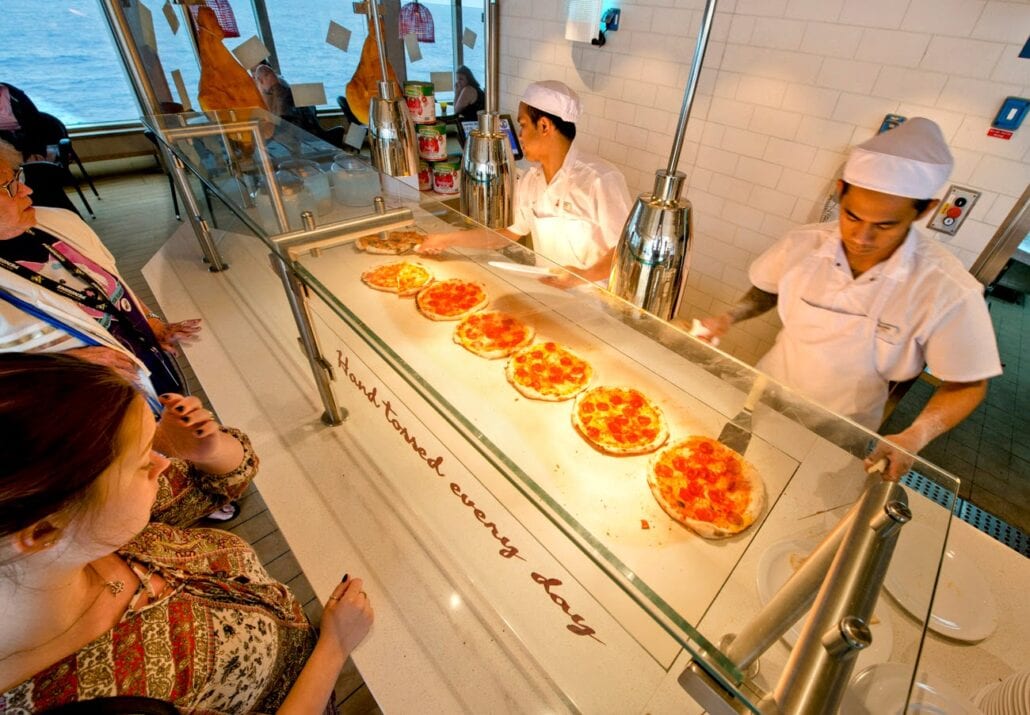 Two chefs in white uniforms and hats prepare and serve several pizzas behind a glass counter to customers, embodying the Carnival Cruise Line spirit. The counter displays the phrase "hand tossed every day" in red lettering as late-night pizza enthusiasts observe the process from the other side.