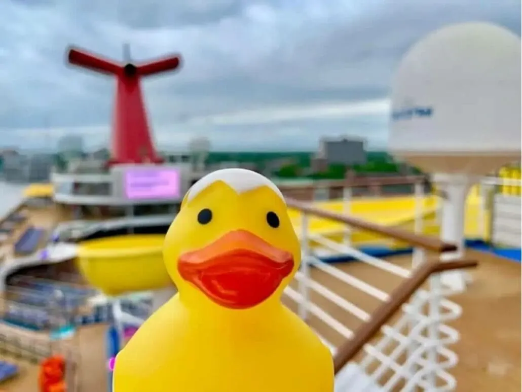 A rubber duck mascot is positioned on the deck of a cruise ship, with the ship's funnel visible in the background. The cloudy sky and some blurred elements create a playful atmosphere reminiscent of carnival fun.