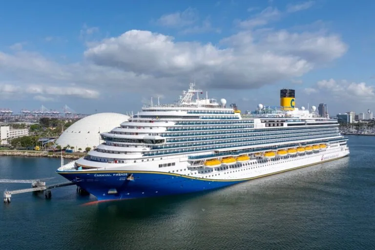 A large Carnival cruise ship docked at a port under a partly cloudy sky. The vessel is white with a blue hull and yellow lifeboats. Several decks are visible, with the New York skyline and its iconic round buildings in the background, offering exciting 2026-2027 options for travelers.