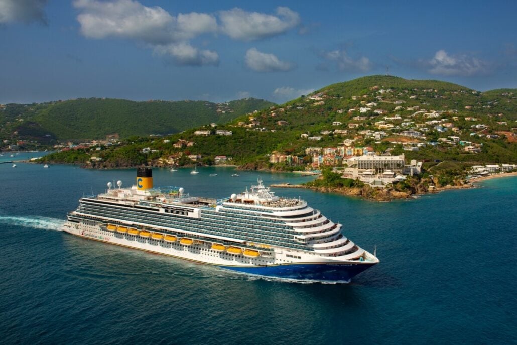 A large Carnival cruise ship sails through a calm blue sea near a lush, green island with scattered buildings. The sky is partly cloudy, and the landscape suggests a tropical or coastal setting, hinting at exciting 2026-2027 options for exploration.