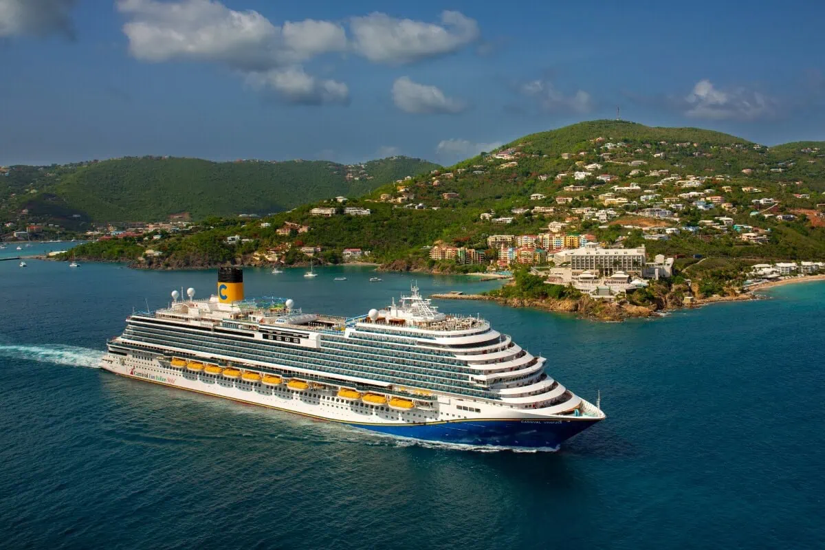 A large Carnival cruise ship sails through a calm blue sea near a lush, green island with scattered buildings. The sky is partly cloudy, and the landscape suggests a tropical or coastal setting, hinting at exciting 2026-2027 options for exploration.