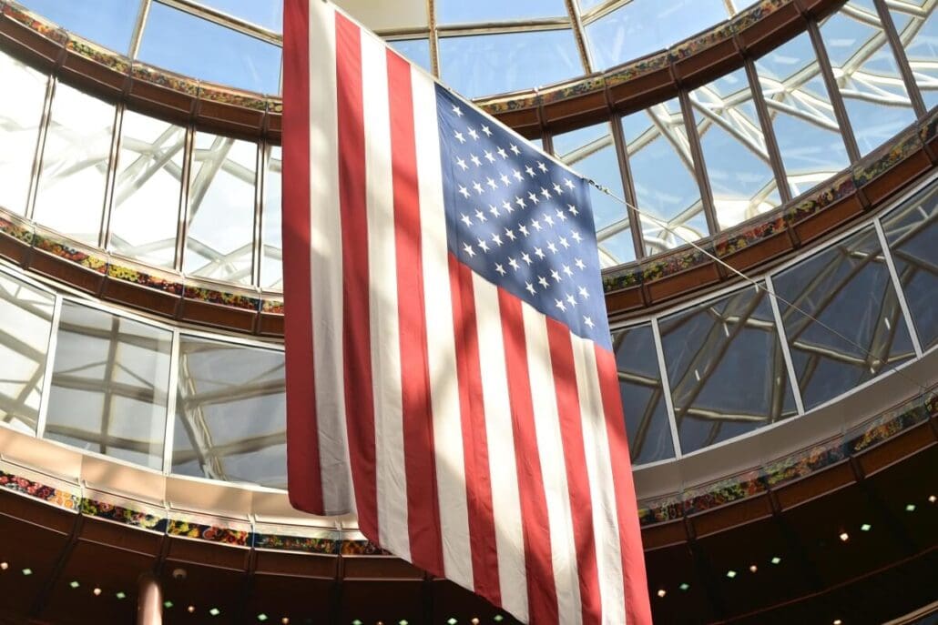 A large American flag hangs indoors from the ceiling of a circular, glass-domed structure. Sunlight filters through the glass, illuminating the flag in anticipation of America's 250th Birthday.
