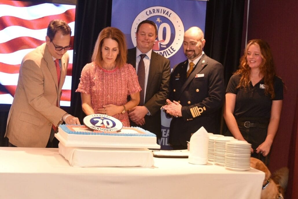 A group of five people stand around a 20th-anniversary cake at a festive carnival event. In Jacksonville, a woman in a pink dress is cutting the cake. Behind them is a banner with a stylized American flag and the number 20, while plates and napkins are neatly arranged on the table.