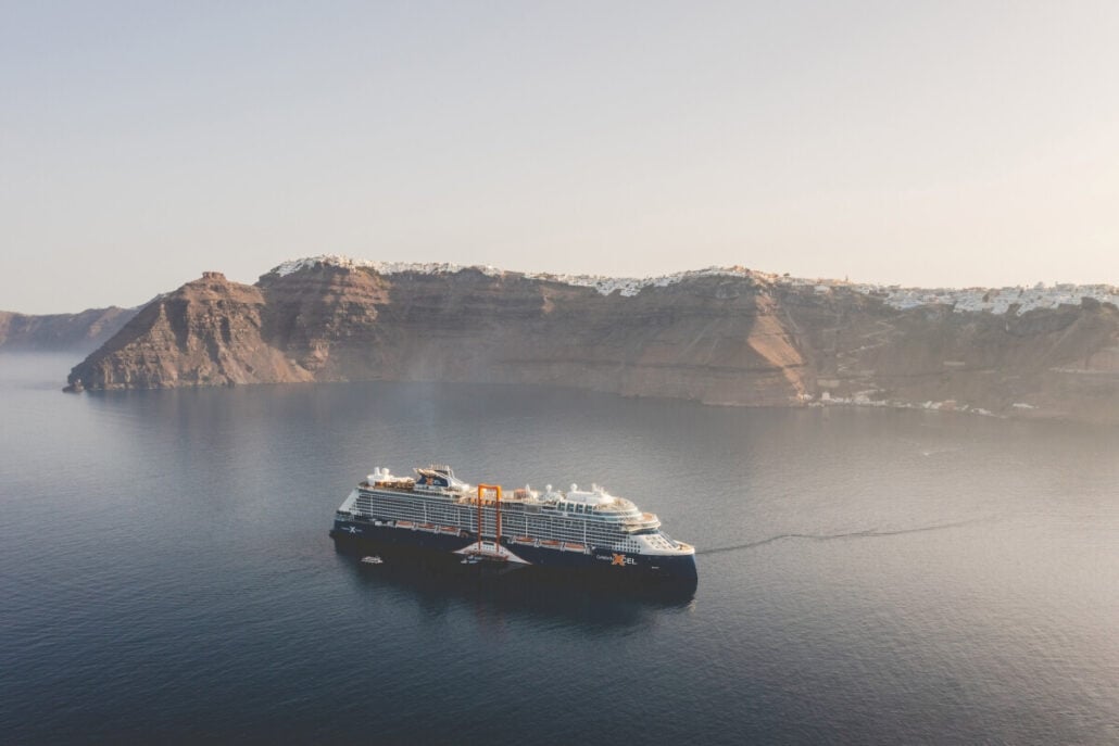 A large cruise ship, reminiscent of the new cruise ships launching in 2025, sails calmly near a rugged coastline. Cliffs rise dramatically in the background with small white structures perched atop. The sky is clear, and the soft lighting suggests early morning or late afternoon.