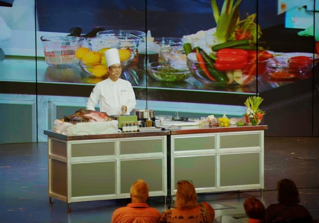 A chef in a white uniform and hat stands behind a counter filled with vegetables and spices, embodying the essence of Holland America's regionally inspired cooking shows. The kitchen setup is displayed on a large screen, while several people sit in front, captivated by the presentation.