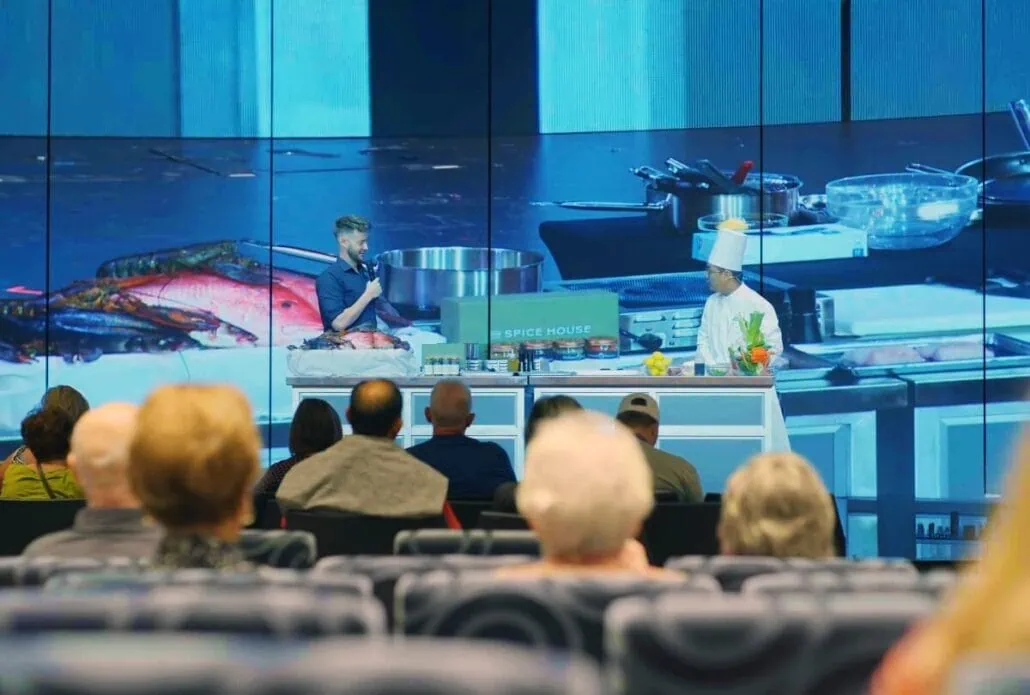 A chef and a man engage in a cooking demonstration on stage, showcasing regionally inspired cooking. Surrounded by utensils and ingredients, they capture the audience's attention in a theater setting. A large screen behind them displays close-ups of seafood and kitchen equipment, reminiscent of popular cooking shows.