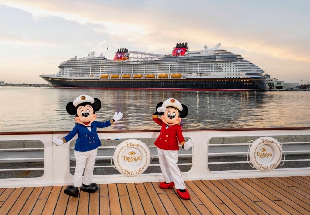 Two mascots dressed as sailors stand on the deck of a ship labeled "Disney Treasure Cruise Line." In the background, Disney’s newest ship departs from Port Canaveral against a stunning sunset.