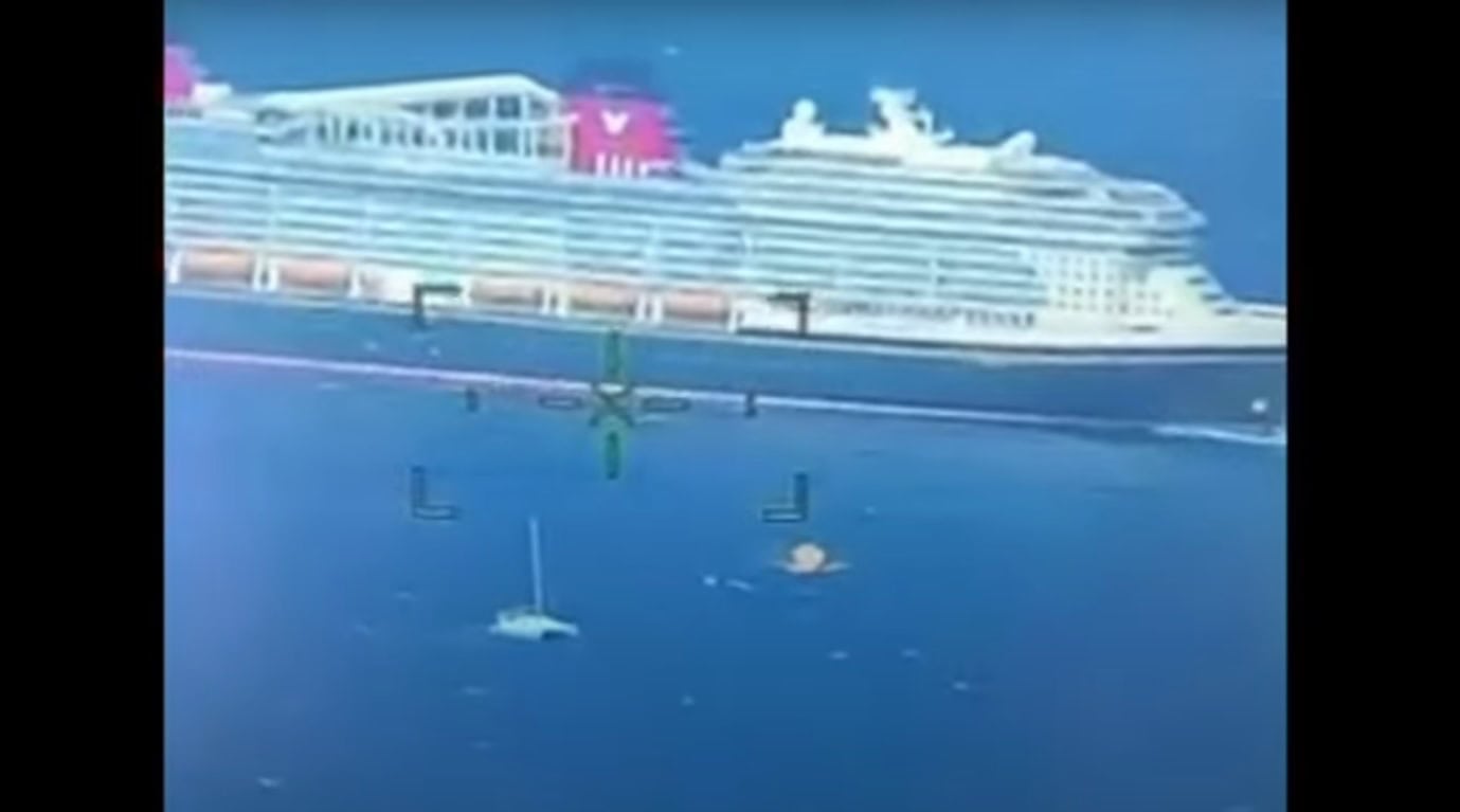 An aerial view captures a massive Disney ship cruising the calm ocean, with a smaller sailing vessel nearby. Crosshairs overlay the image, suggesting it's taken from a surveillance helicopter during a rescue mission. The sea is tranquil, and the sky is clear.