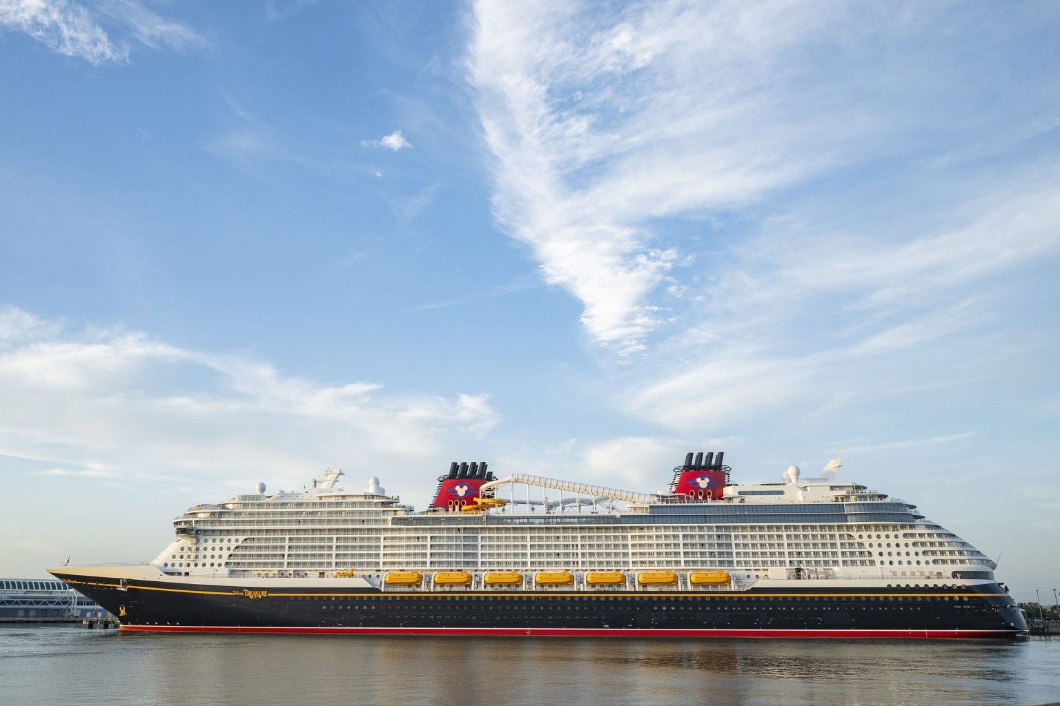 Disney's newest ship gracefully floats on calm water under a blue sky with scattered clouds, ready to depart from Port Canaveral. The vessel features prominent red and black funnels and multiple decks lined with yellow lifeboats, promising an unforgettable journey.