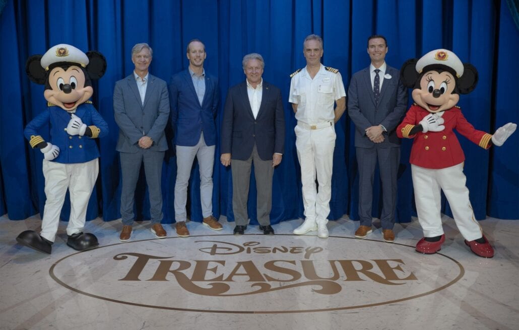 People stand on a stage with a "Disney Treasure" logo on the floor. Two individuals are dressed as characters in blue and red uniforms with sailor hats. Four other men in business and naval attire stand in front of a blue curtain at Port Canaveral, celebrating Disney’s newest ship.