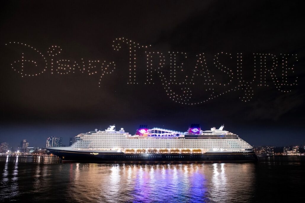 A large cruise ship is illuminated on the Hudson River at night. Above, a dazzling light show forms the words "Disney Treasure" in the sky, while city buildings and their reflections twinkle in the background.