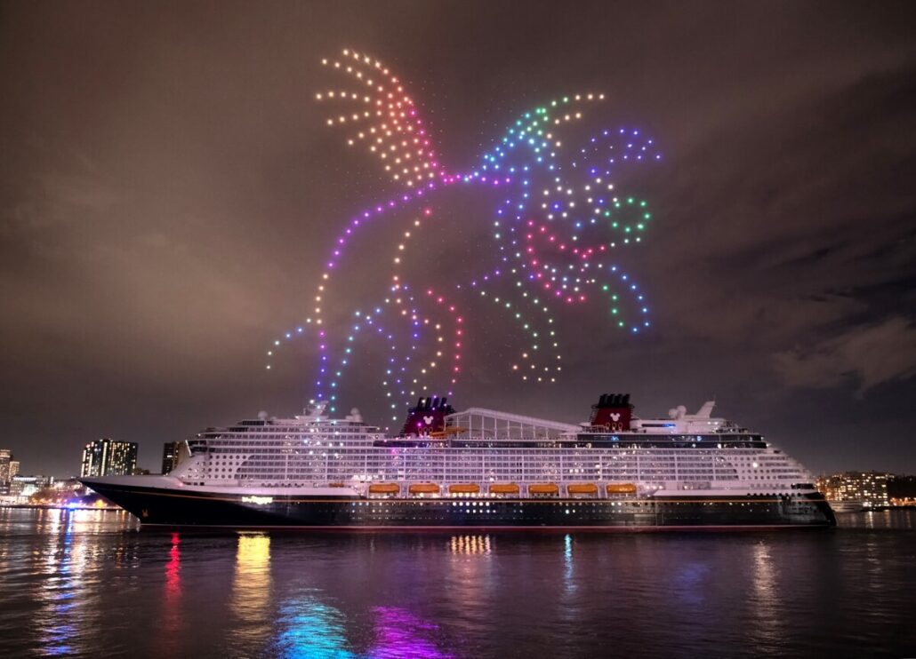A large cruise ship is docked at night along the Hudson River, with a dazzling light show featuring a winged creature, possibly a dragon, illuminating the sky. The Disney Treasure adds magic to the scene as city lights twinkle in the background.