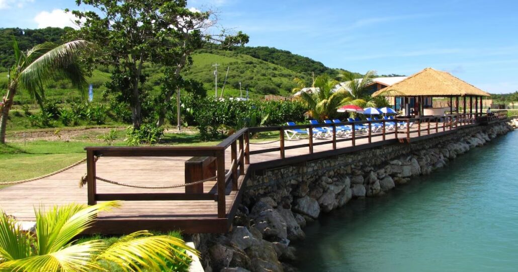 A wooden walkway bordered by a rope leads to a thatched-roof building near the water, reminiscent of Montego Bay. The area is surrounded by lush greenery and palm trees, with blue lounge chairs visible under umbrellas. The sky is clear and sunny.