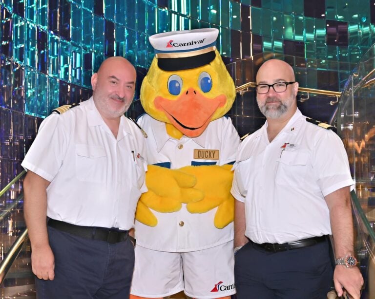 Two uniformed men stand on either side of a person in a duck mascot costume, wearing a sailor hat labeled "Ducky," as if welcoming visitors to the carnival-themed cruise. They pose indoors, surrounded by a shiny, colorful backdrop reminiscent of vibrant festivities.