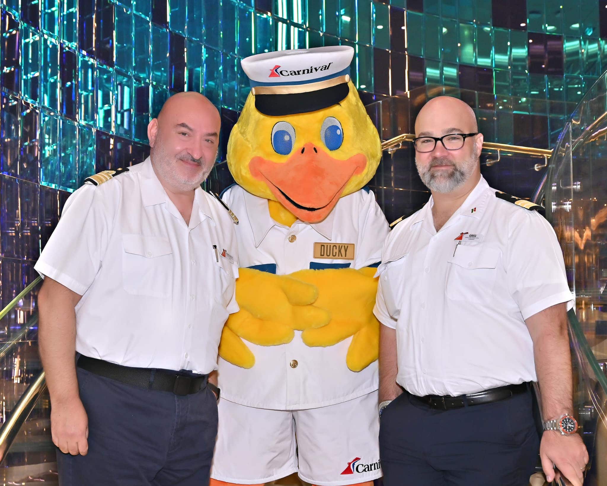 Two uniformed men stand on either side of a person in a duck mascot costume, wearing a sailor hat labeled "Ducky," as if welcoming visitors to the carnival-themed cruise. They pose indoors, surrounded by a shiny, colorful backdrop reminiscent of vibrant festivities.