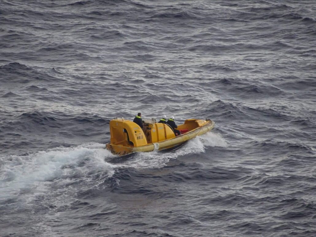 A yellow rescue boat is navigating through choppy waters with three people on board, all wearing life jackets and helmets. The boat, possibly part of a Royal Caribbean mission, is moving to the right against a backdrop of gray waves, searching for a stranded family.