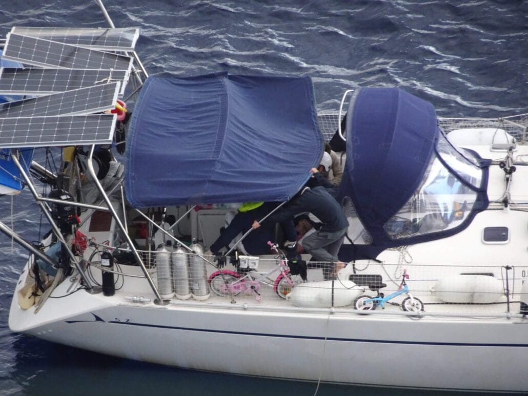 A sailboat on the water with two bicycles secured on deck showcases a self-sufficient lifestyle. Equipped with solar panels and covered areas, it hosts two people engaged in activities as they navigate the serene ocean, perhaps dreaming of distant destinations like Africa.