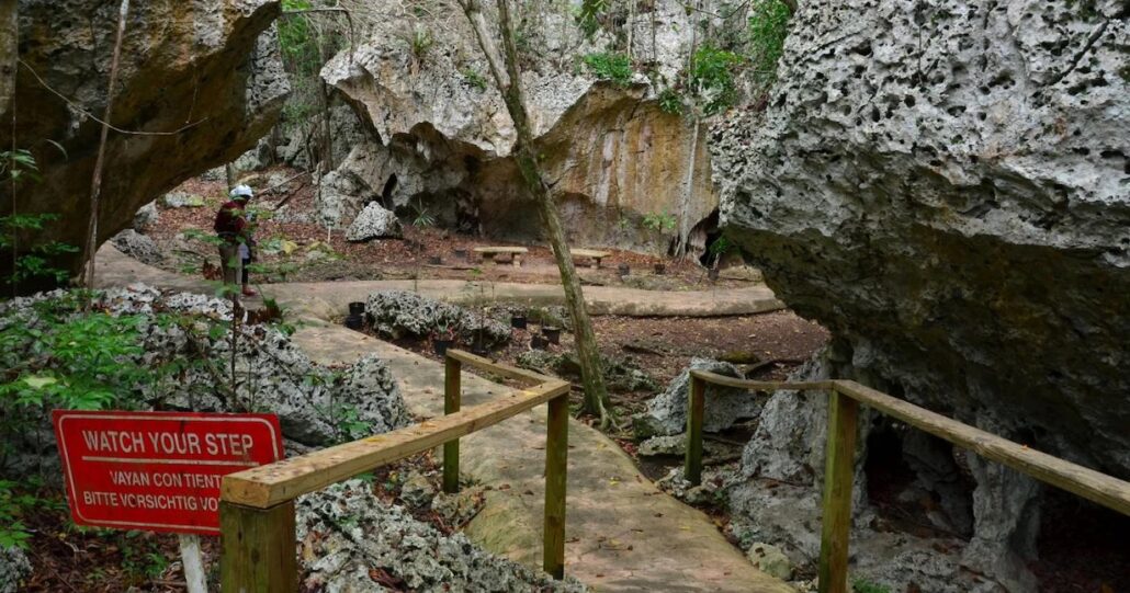 A rocky path winds through a forested area reminiscent of Montego Bay, with large boulders and a wooden railing lining part of the way. A red sign warns, "Watch your step." Trees and lush greenery provide a scenic backdrop as two people wearing helmets traverse the trail.