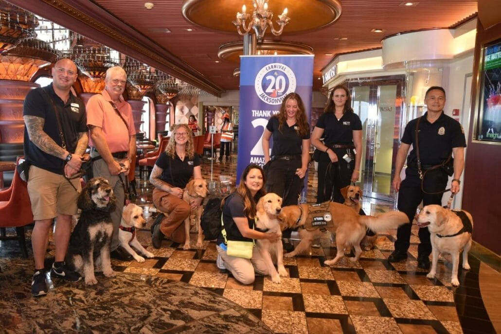 In Jacksonville, a group of people and dogs pose in an inviting indoor setting with wood-paneled decor. A banner proudly reads "Carnival Cruise Line 20 Amazing Years!" Some dogs wear harnesses or vests, highlighting their role as service animals.