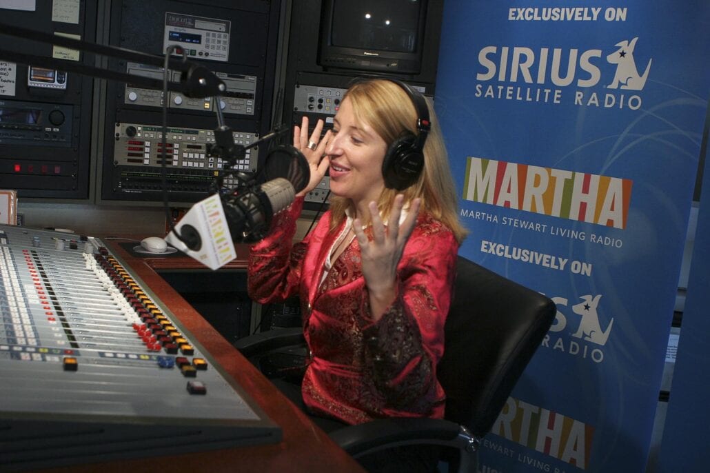 A woman wearing headphones, seated at a radio studio desk with a microphone and mixing board, gestures animatedly. Behind her are banners for Sirius Satellite Radio and Martha Stewart Living Radio, reflecting her career passion and happiness.