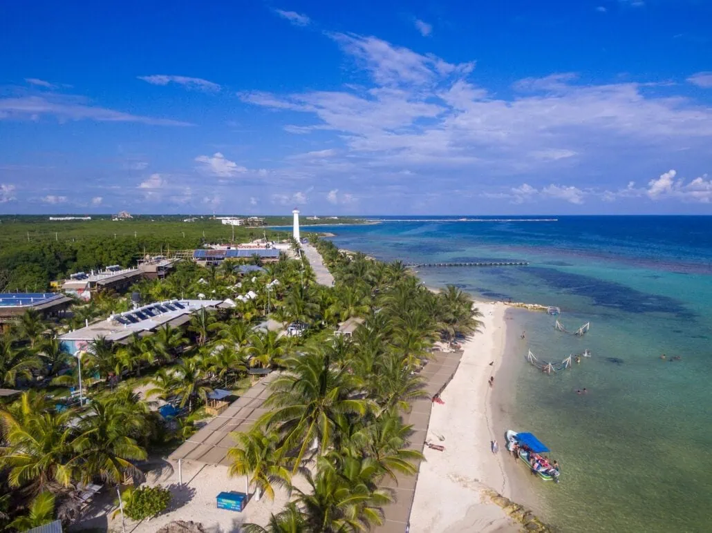 Aerial view of Costa Maya's beach with white sand and clear blue water, bordered by palm trees and a few structures. Cruisers are swimming and relaxing on the shore. A boat is anchored near the beach, and a lighthouse stands in the distance.