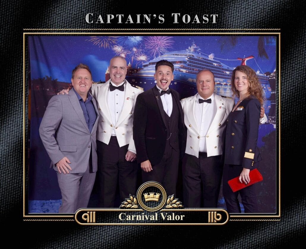 A group of five people, including Fleet Cruise Director Mike Pack, are dressed semi-formally and formally, posing together in front of a cruise ship backdrop. The image is framed with the text "Captain's Toast" and "Carnival Valor" at the bottom.