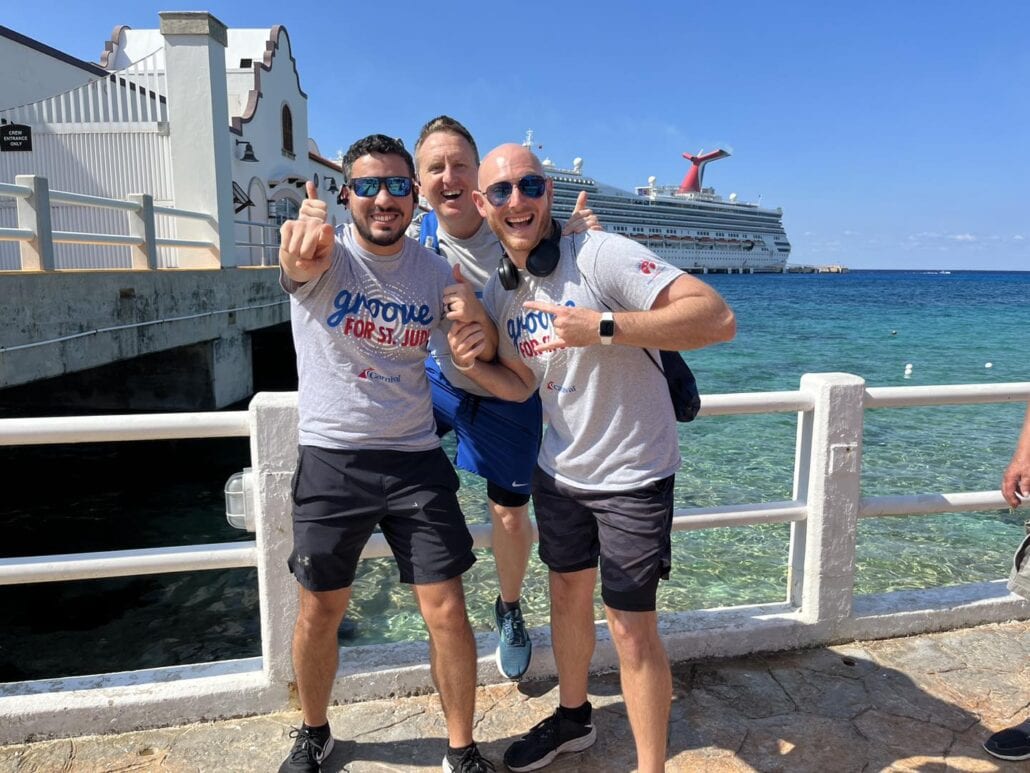 Three men smiling by the waterfront with a large Carnival cruise ship in the background. Two wear matching gray shirts that read "Gigique for St. Jude." The Fleet Cruise Director, Mike Pack, sporting sunglasses, points at the camera as the sky remains clear and sunny.