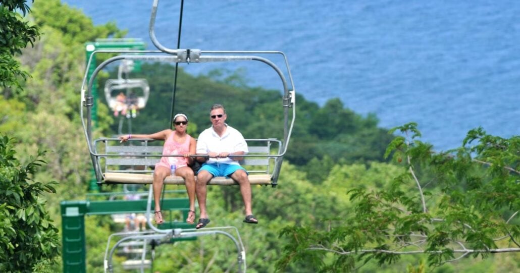 A man and a woman are seated on a ski lift overlooking the lush, green landscape of Montego Bay, with the serene blue water in the background. The man wears a white shirt and blue shorts, while the woman dons a pink outfit and sunglasses.