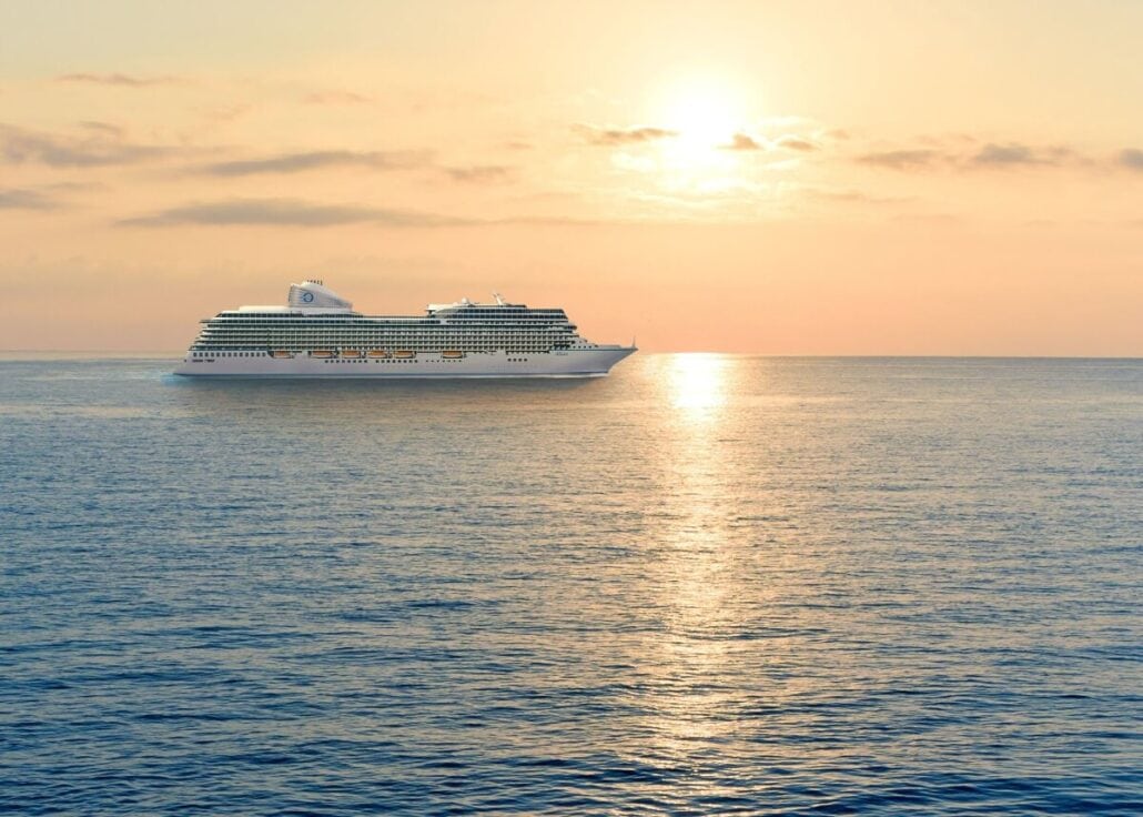 A large cruise ship, reminiscent of the new cruise ships in 2025, sails on a calm ocean under a sunset sky. The sun is partially obscured by light clouds, casting a warm glow on the water. The ship is positioned slightly to the left of center, heading toward the horizon.