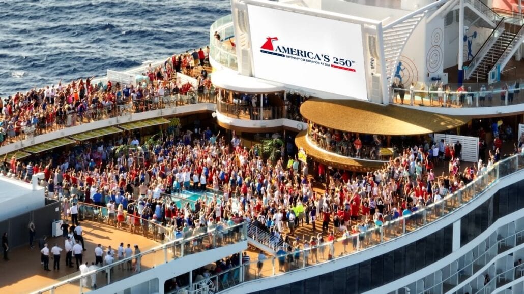 A large crowd gathers on the deck of a Carnival cruise ship. A prominent sign displaying "America's 250th Birthday" is visible. The vessel is at sea, and people appear to be attending a festive celebration.