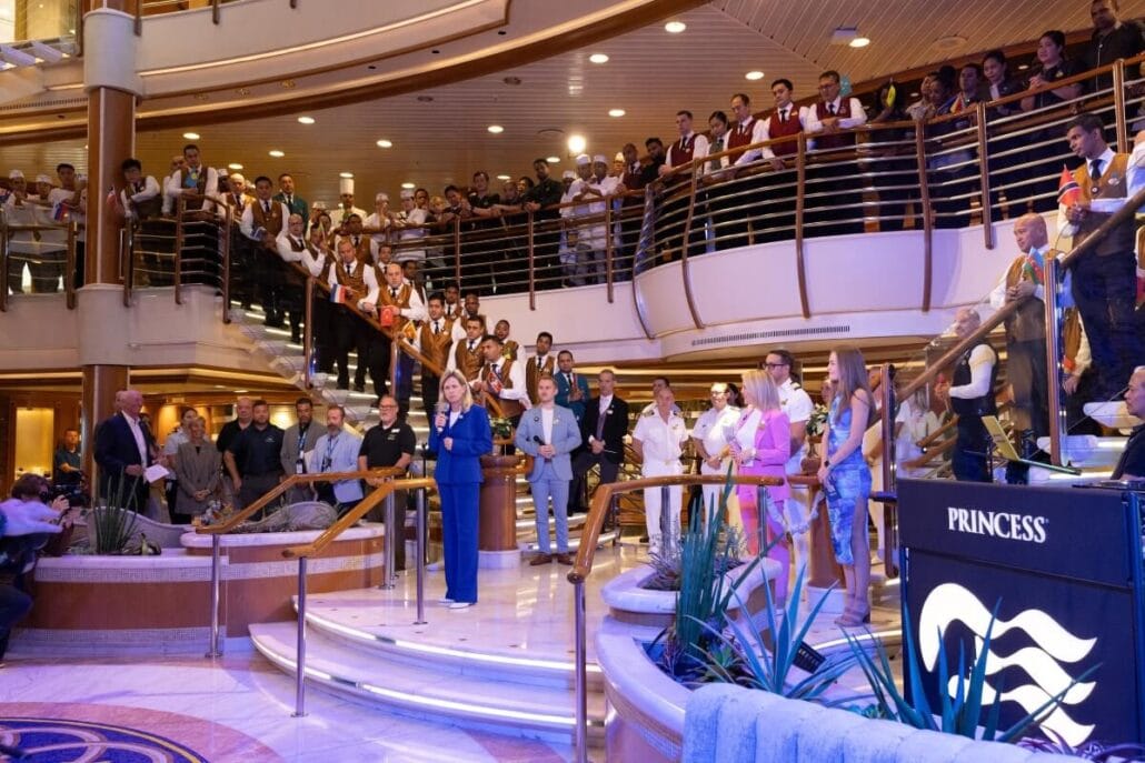 A person is addressing a gathered crowd on the multi-level staircase in a cruise ship's atrium. Several people, some in uniform, are listening attentively. A screen displays "Princess" with a wave logo, reminiscent of scenes from "The Love Boat" as it sets sail from Port Canaveral this Winter Season.