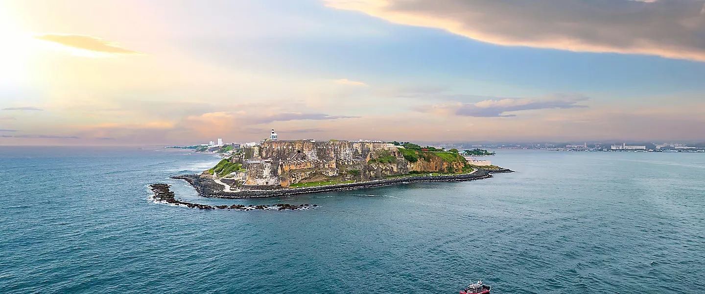 View of a small island with rugged cliffs surrounded by the ocean, possibly near Puerto Rico. The island features a historic-looking fortress and a lighthouse, with large ships occasionally making port calls at the city visible in the distant background. The sky is partly cloudy with a soft sunset glow.