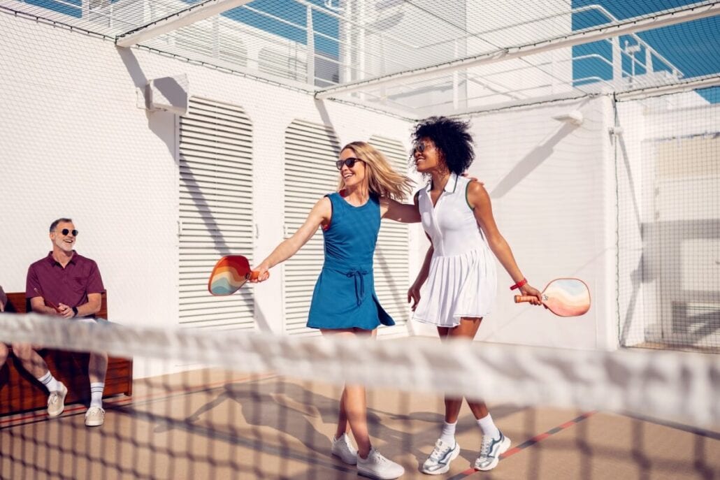 Two women in athletic dresses play padel on an outdoor court with a netted enclosure, enjoying the cognitive benefits of this engaging sport. They hold paddles and move dynamically, while a man in sunglasses sits on a bench, cruising through the sunny day as he watches them.
