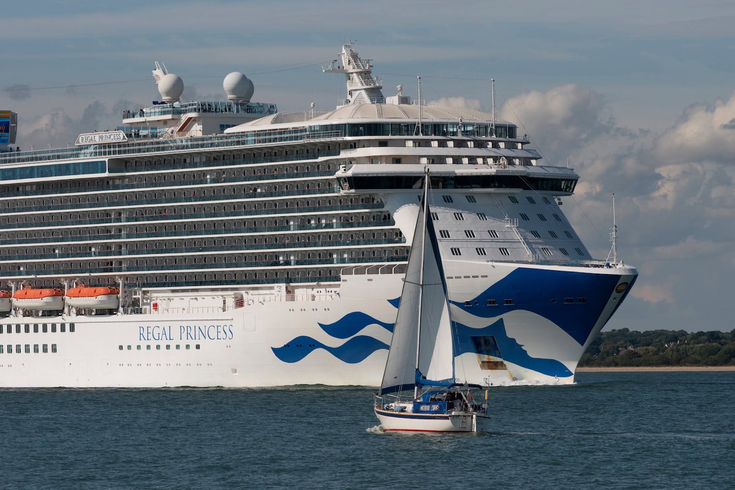The impressive "Regal Princess" from Princess Cruises gracefully glides alongside a small sailboat in open water. With the sky partly cloudy and land visible in the background, passengers dream of future cruise adventures amid the possibilities of their Future Cruise Credit.