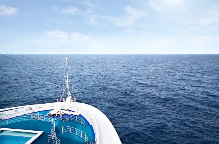 The image captures a traveler's paradise with the bow of a cruise ship slicing through a calm ocean. Luxurious cruise ship rooms overlook the inviting swimming pool and sun-drenched deck chairs, set against a clear, sunny sky.