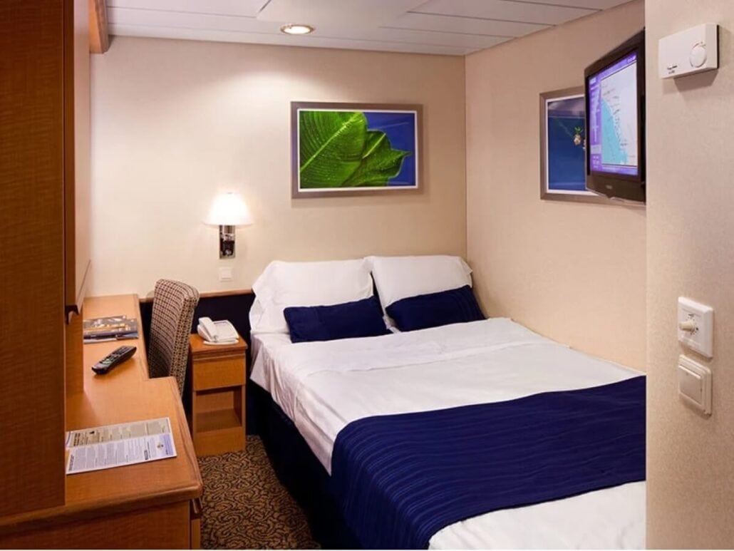 A small, neatly arranged cruise ship cabin offers travelers a best-in-class experience with a bed draped in white sheets and a dark blue throw. The room includes a wall-mounted TV, a desk with chair, decorative artwork, and soft lighting that beautifully illuminates the space.