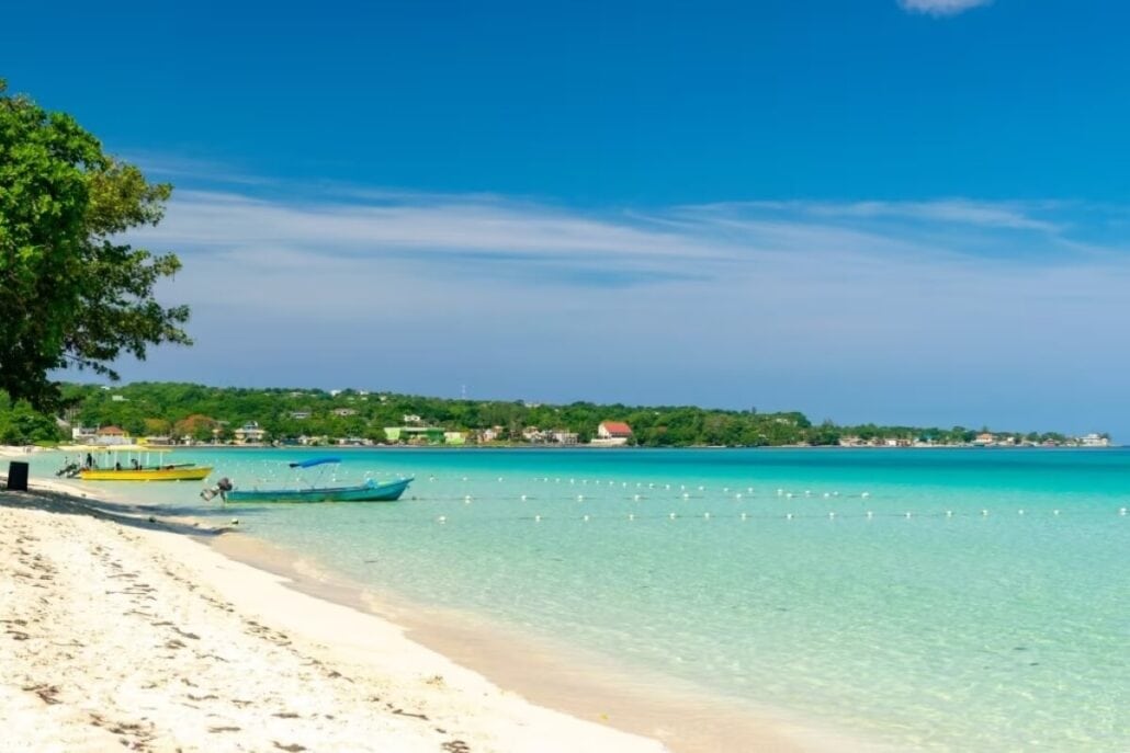 A clear, sunny day at a tropical beach in Montego Bay features turquoise waters and white sand. A few small boats are anchored near the shore, with lush green trees lining the coast. In the distance, buildings and vibrant greenery complete this picturesque scene.