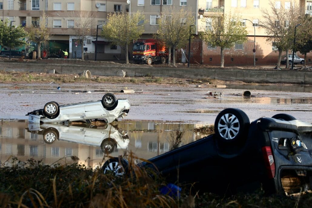 Two overturned cars lie partially submerged in the flood-ravaged area of Valencia, near residential buildings. A fire truck and emergency personnel can be seen in the background, with scattered debris floating in the water. Trees line the sidewalk along the buildings, mirroring Spain's resilient spirit.