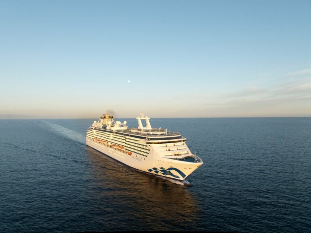 A large Princess Cruises ship sails under a clear blue sky on a calm ocean. The vessel, part of their 131-day sailing itinerary covering 60 destinations, moves away from the camera, leaving a trail in the water. The horizon is visible in the distance, with a full moon gracing the sky.