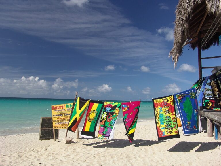 Colorful beach towels and flags hang from a wooden structure on a sandy Montego Bay beach. The background features a turquoise ocean and partly cloudy sky, with a sign displaying various text on the left.