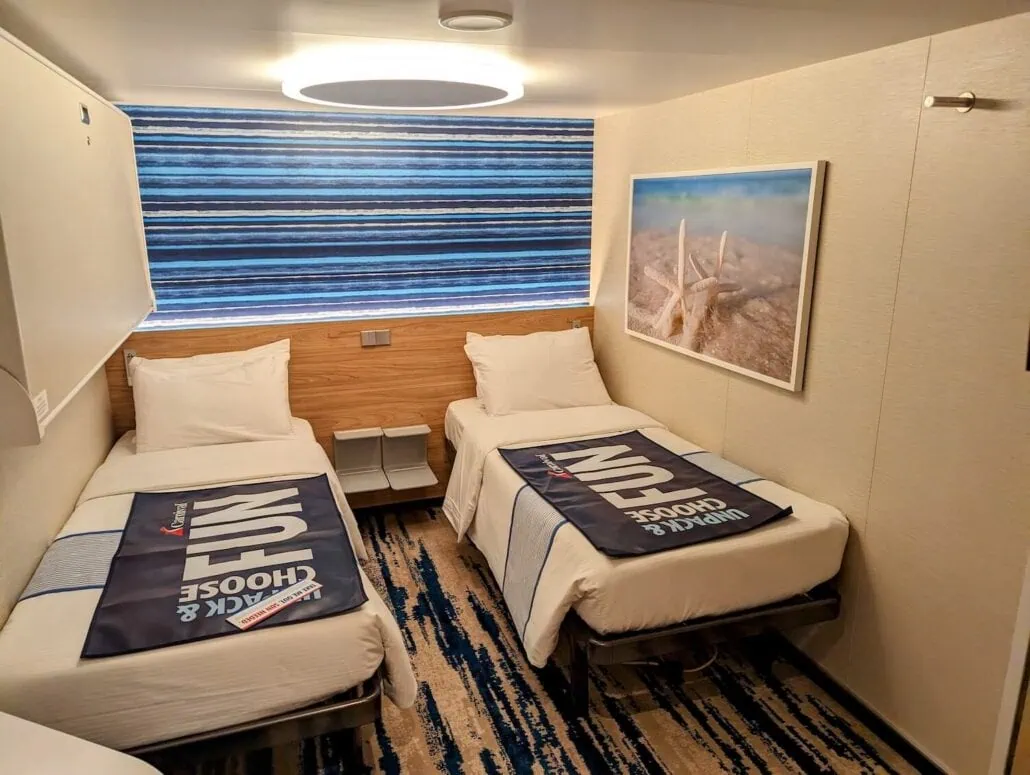 A cozy Carnival Cruise Line cabin room features two single beds with white linens and decorative towels, a framed picture of starfish on the wall, and a blue and white striped window covering. The carpeted floor, in tones of blue and beige, adds to the nautical charm.
