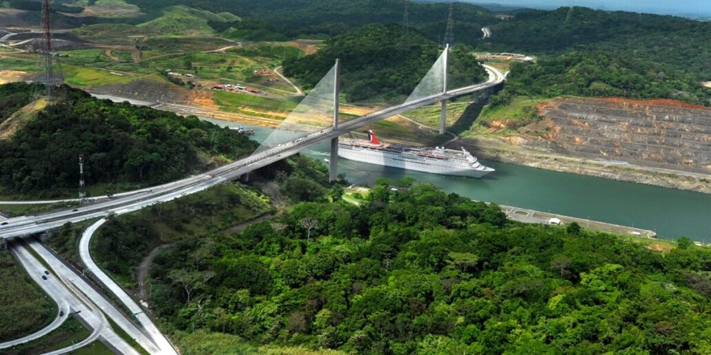 Aerial view of a Carnival cruise ship passing under a tall, modern cable-stayed bridge spanning a wide canal. The surrounding area features lush green forests and roadways. Hills and construction sites are visible in the background, making it clear why passport-required Panama visits are unforgettable experiences.