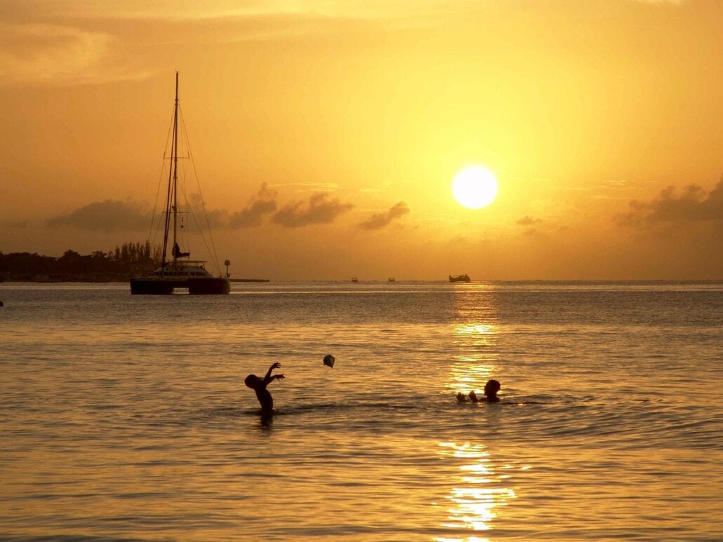 Two people are playing in the water at sunset in Montego Bay. A sailboat is anchored in the distance, and the golden sky reflects on the calm sea, creating a serene atmosphere.