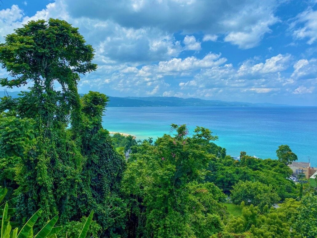 Lush green trees in the foreground frame a breathtaking view of the turquoise sea, reminiscent of Montego Bay, with distant mountains under a partly cloudy blue sky.