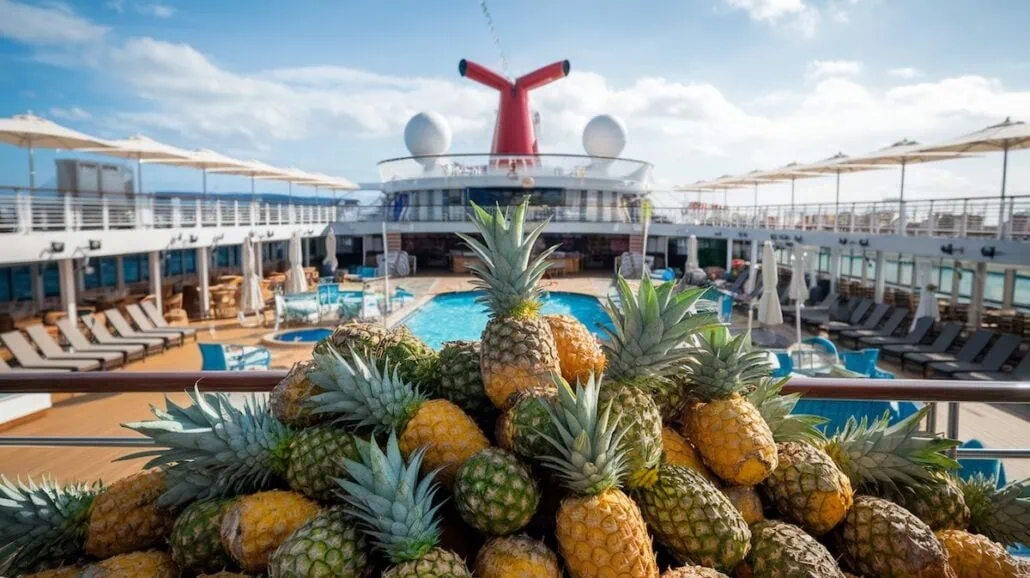 A pile of upside-down pineapples is placed on a Carnival Cruise Line deck, with a swimming pool and lounge chairs visible in the background. The ship's distinctive red funnel and two white domes are centered in the image under a partly cloudy sky.
