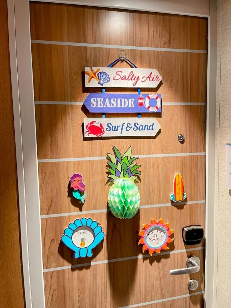 A wooden cabin door adorned with beach-themed signs and items showcases colorful cutouts of a starfish, shell, surfboard, sun, and mermaid. An upside-down pineapple adds a whimsical touch alongside signs reading "Salty Air," "Seaside," and "Surf & Sand.