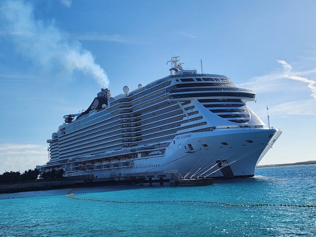 A large cruise ship docked at a port in turquoise sea water under a clear blue sky. The vessel has multiple decks with visible balconies and a smoking funnel at the top, hinting at minor power issues. A distant shoreline and a bright sunny day complete the scene.
