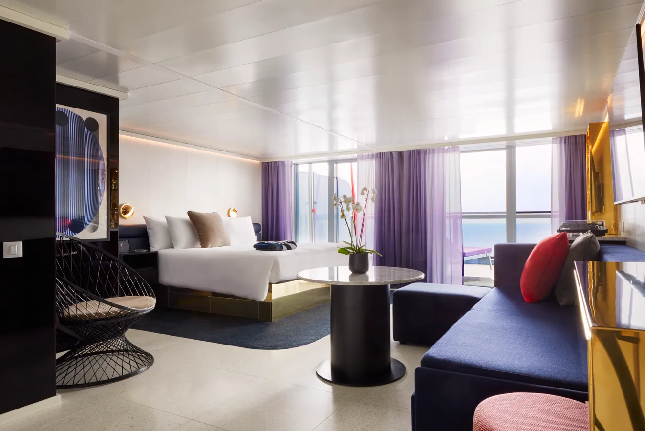A modern hotel room aboard the Scarlet Lady returns to service with a large bed, a seating area featuring a blue sofa and a round black table, and a balcony offering an ocean view. The room boasts purple curtains, artistic decor, and a potted plant on the table after its dry dock upgrades.