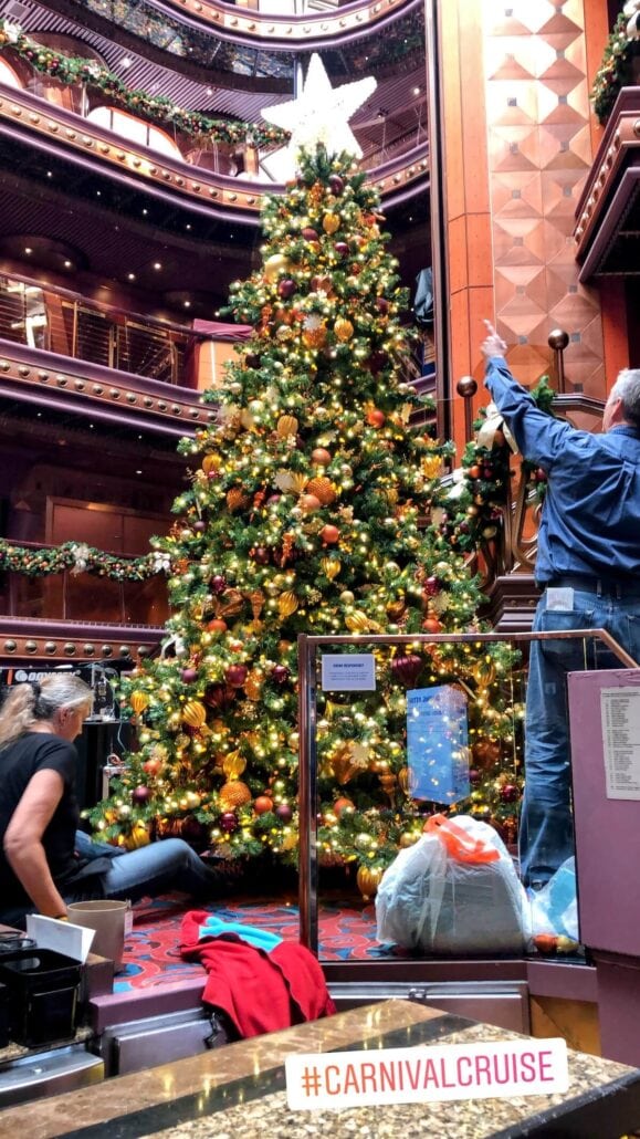 A large, decorated Christmas tree stands in the center of a cruise ship's atrium, transforming it into a holiday wonderland. Two people arrange decorations amidst festive garlands and a spacious interior. A #CarnivalCruise tag is visible at the bottom.